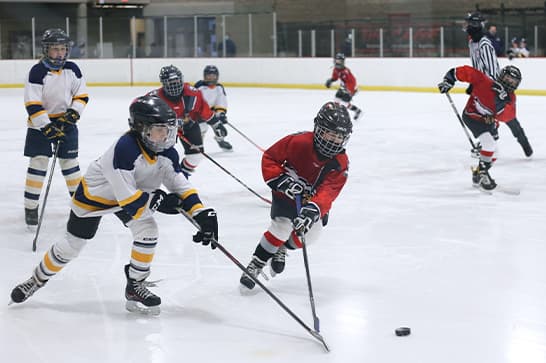 Youth hockey game on an ice rink; players in red and white jerseys chase the puck as a defender closes in, with a referee visible in the background.