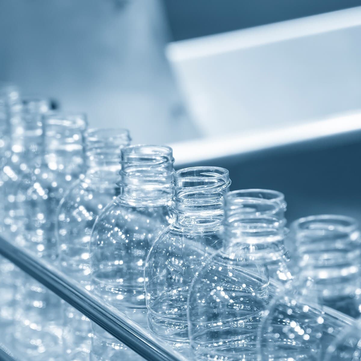 Row of empty glass bottles on a moving conveyor in a blue-toned industrial setting, with light reflecting off their surfaces.