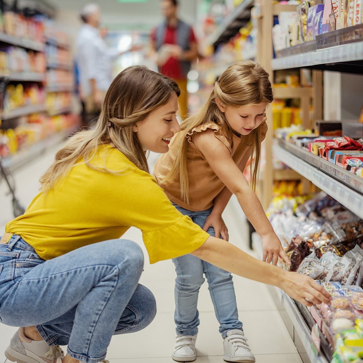 A caregiver and a child crouch in a grocery store aisle, looking at snack packages on a shelf.