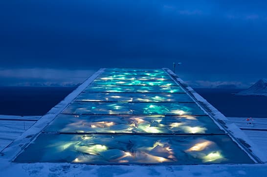 A glowing glass walkway over snow and ice leads toward a distant blue horizon, with multicolored lights beneath the panels.