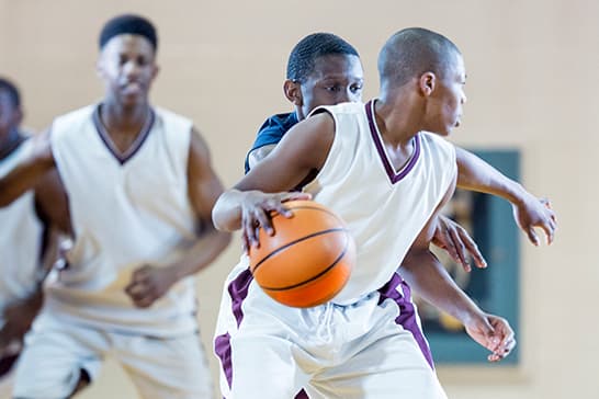 Three players in white and purple uniforms play basketball on an indoor court; the foreground player grips an orange ball while others guard.
