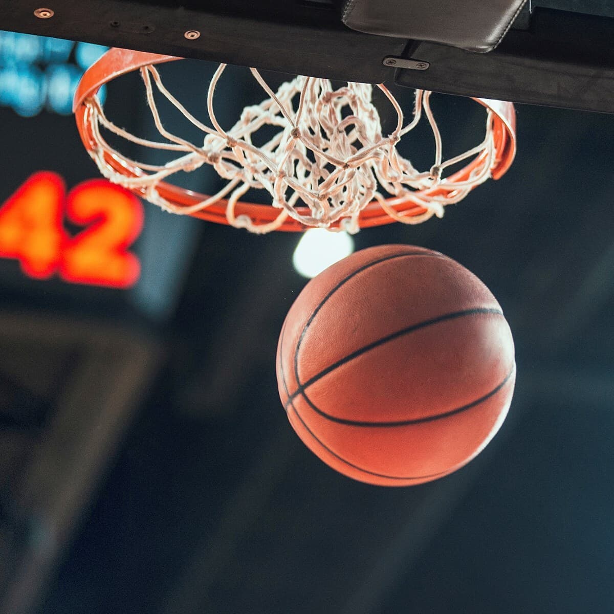 Close-up of a basketball entering the hoop with an orange rim and tangled white net, blurred 42 on a scoreboard in the background.