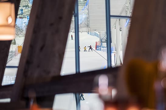 Indoor ice rink seen through dark wooden beams; several skaters on the bright ice with glass walls and bright background.