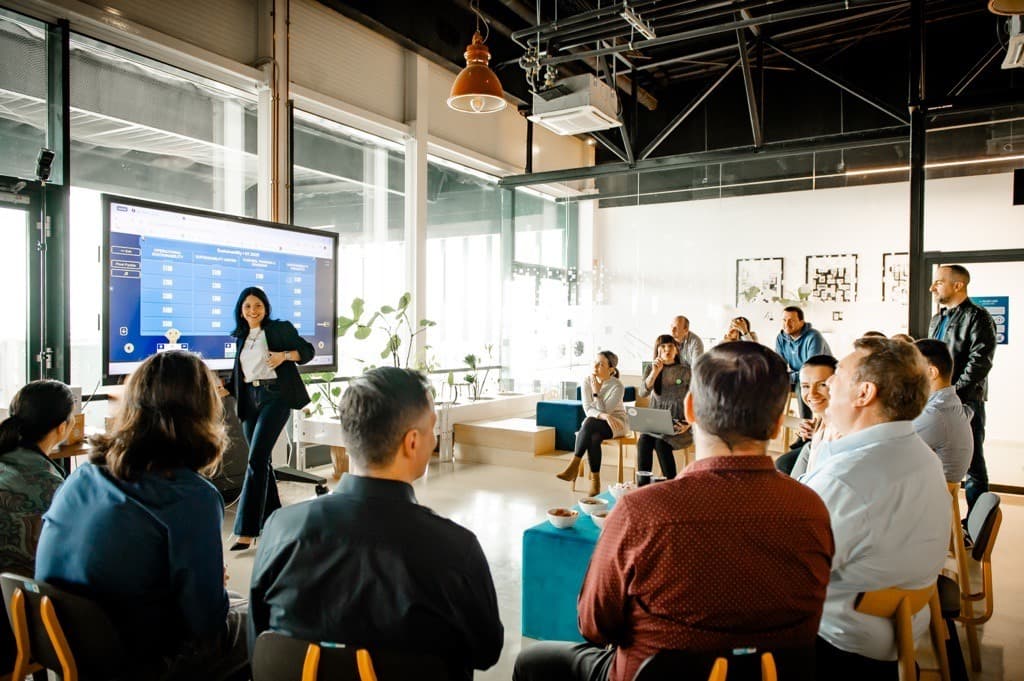A presenter leads a workshop in a bright, modern open office with a large screen; diverse group seated in a circle, listening and smiling.