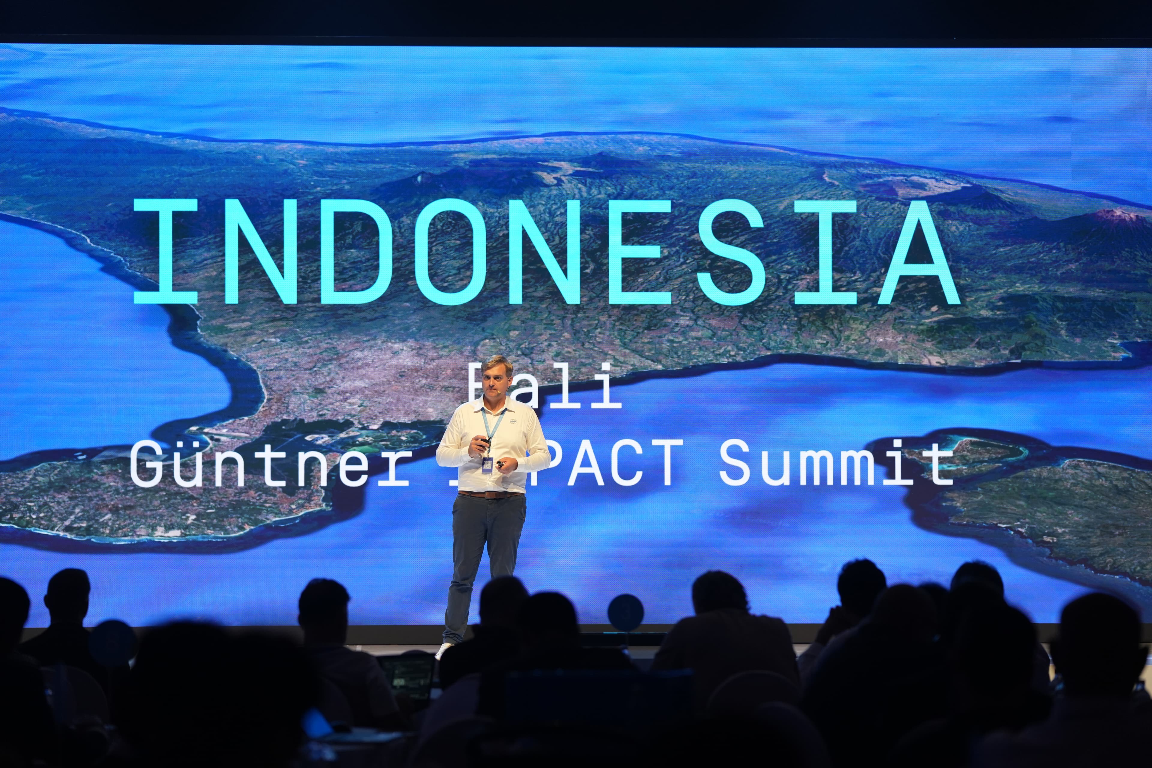 Presenter in a white shirt on stage before a large screen showing a map of Indonesia with turquoise “INDONESIA” text and “Bali”; audience silhouettes in front.