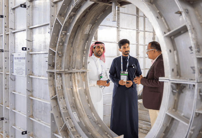 Three men stand inside a large metal industrial frame, discussing while wearing name badges; one in a traditional white robe and headscarf, another in a dark robe, and a third in a blazer.
