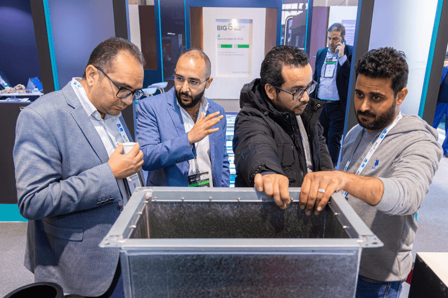 Four men in business casual attire gather around a metallic tank or display at a trade show, one pointing, another examining, all focused on the object.