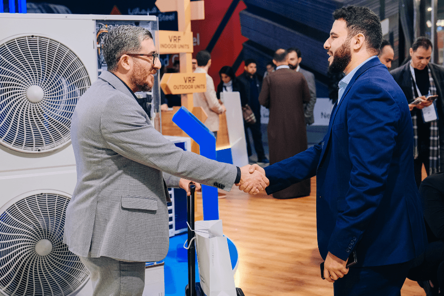 Two men in suits shake hands at a trade show, with large air conditioner units behind them and attendees in the background browsing booths.
