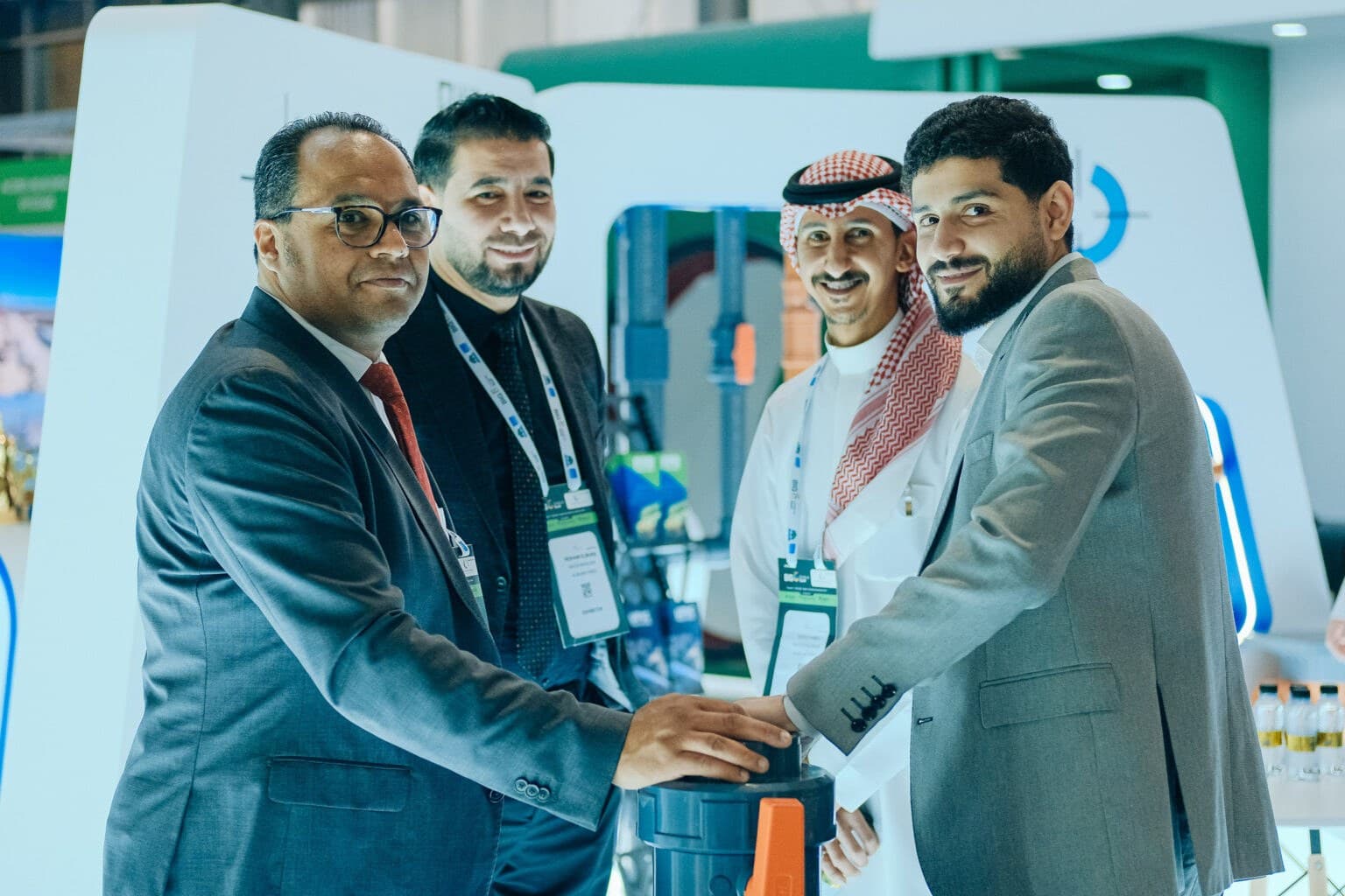 Four diverse professionals in business attire shake hands around a tech exhibit booth, smiling, with lanyards and a large white display in the background.