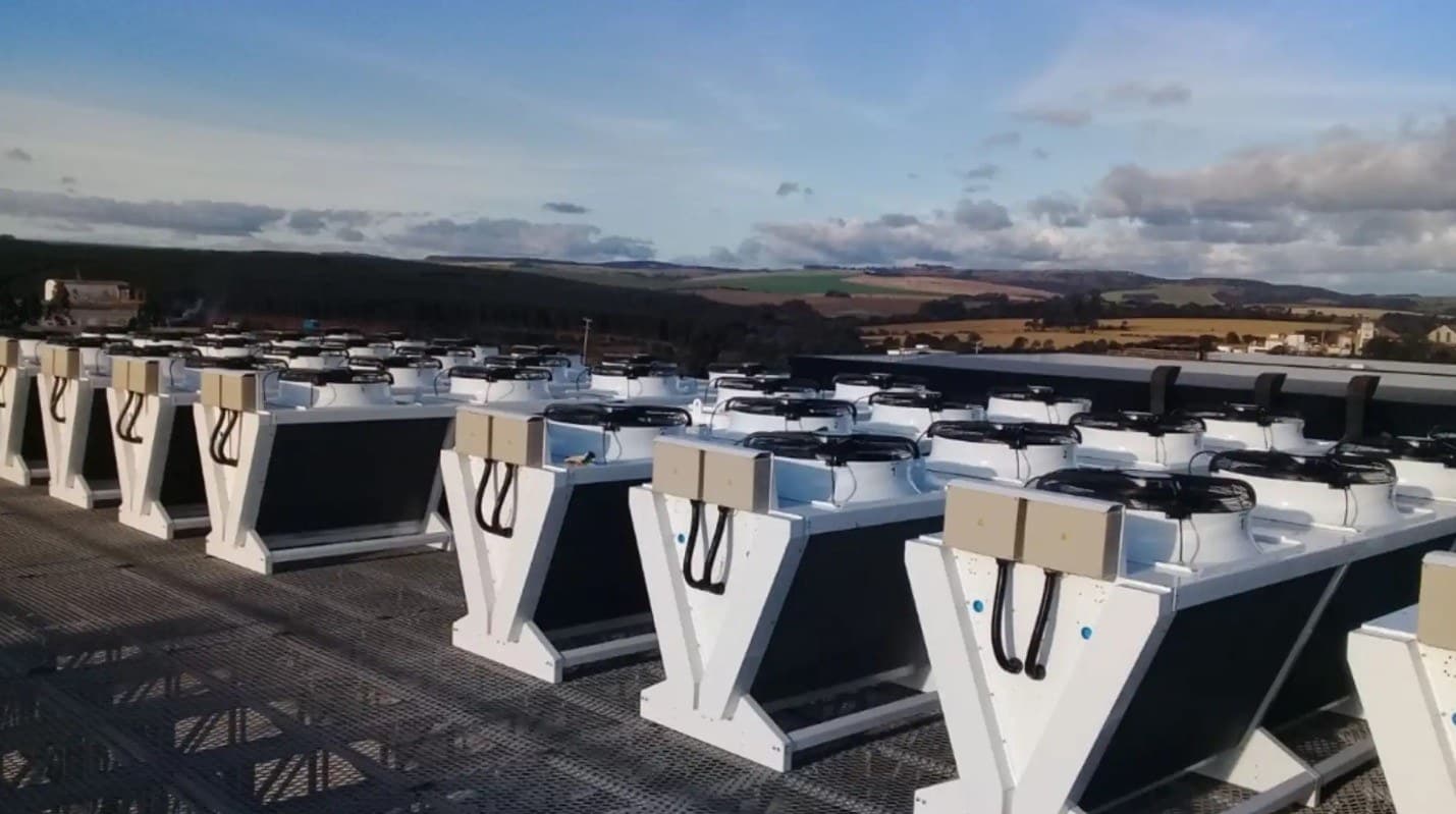 Row of white industrial cooling units with black circular fans on top, on a rooftop, with distant hills and a blue sky.