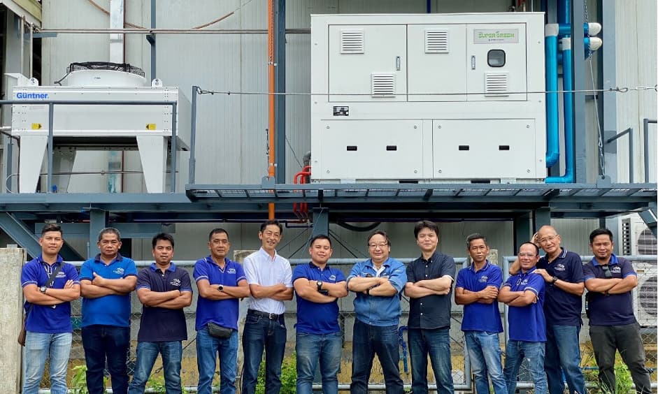 Group of people in blue polo shirts with arms crossed, standing in front of large industrial equipment on an outdoor platform.