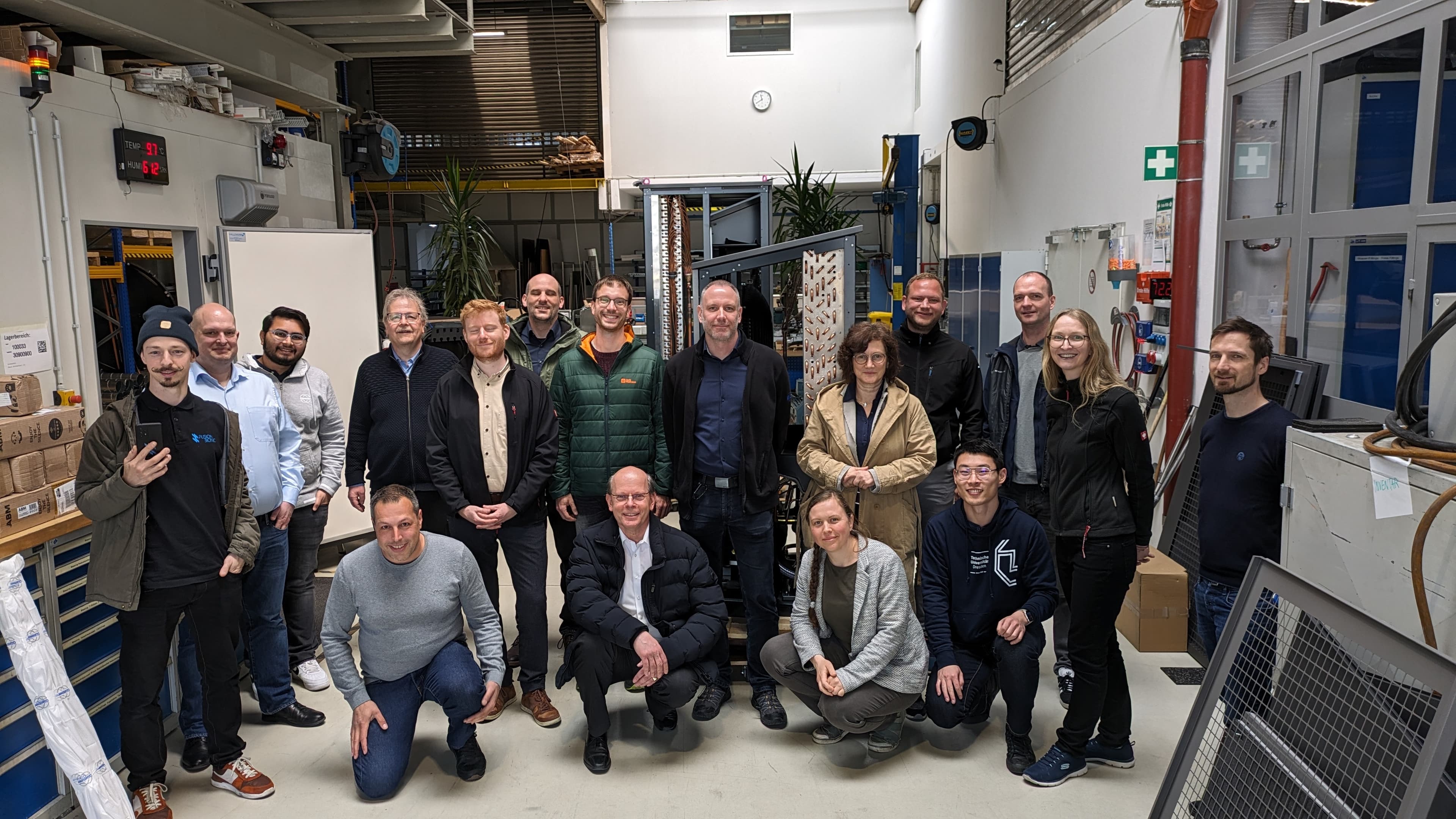 Diverse group of coworkers posing for a photo in a bright industrial workshop with machines, shelving, and safety signs.