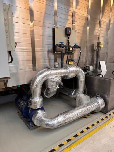 Industrial mechanical room with large insulated silver pipes, blue valves, a pump and gauges, plus a control panel; yellow and black floor safety tape along the edge.