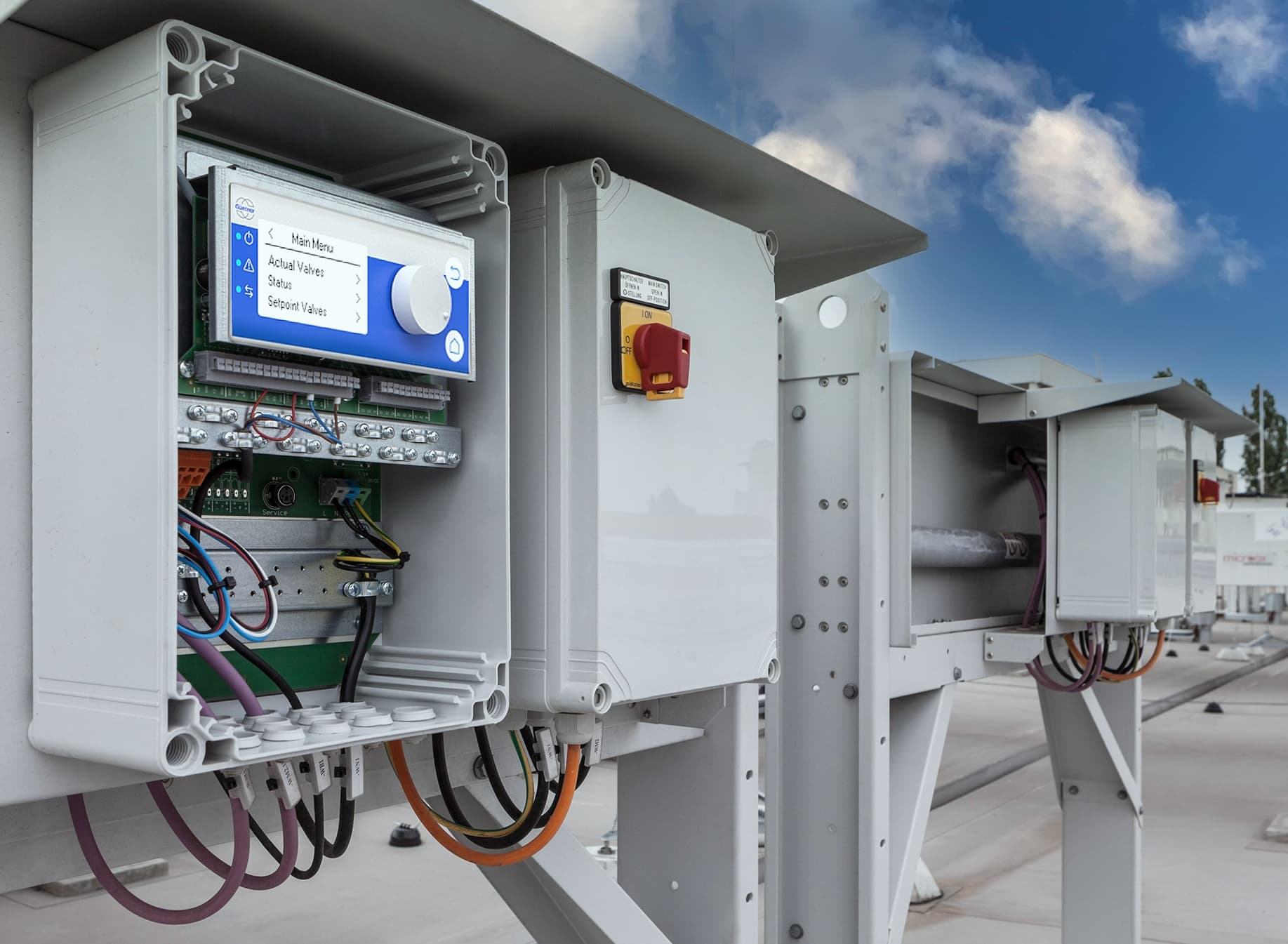 Open industrial control cabinet mounted on a gray metal frame, featuring a blue display panel, circuit boards and colorful wires under a clear blue sky.