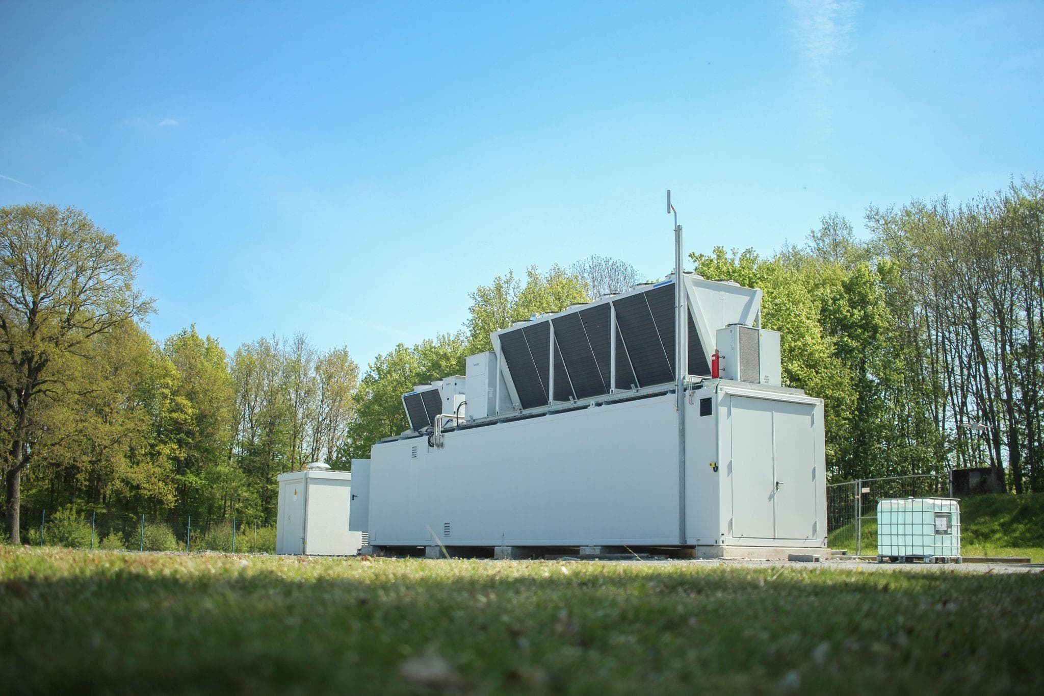 White industrial unit with angled solar panels on top sits on a grassy field with trees and blue sky; a small container and an IBC tote nearby.