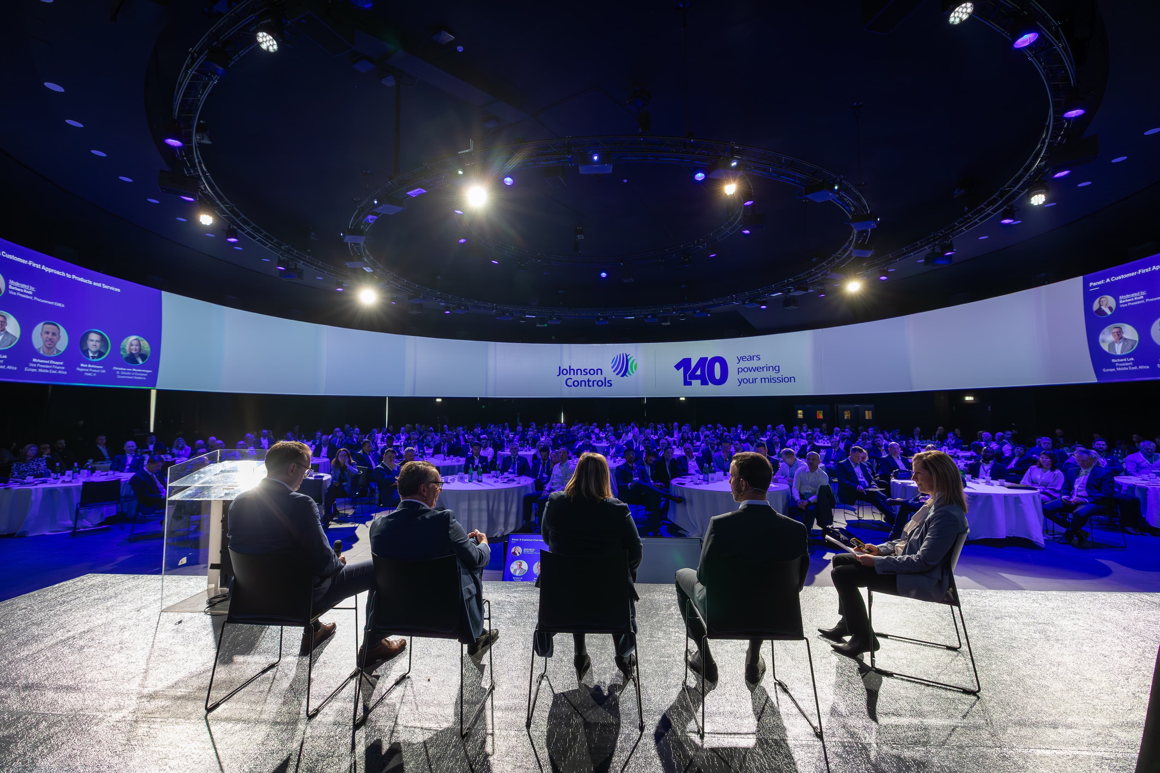Panel of five on stage addressing a large audience at a Johnson Controls event, with a circular lighting rig overhead.