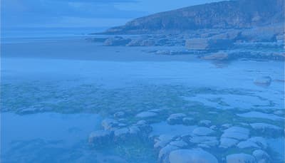 Calm coastal scene with clear blue water over rocks in the foreground and a rocky shoreline extending toward distant hills.
