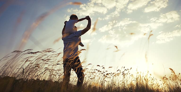 A person carries a child on their shoulders in a sunlit field of tall grass at sunset, with warm golden light.