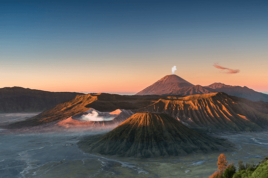 Sunrise over a volcanic landscape with a cone-shaped foreground volcano and a taller smoking volcano in the distance under a blue-orange sky.