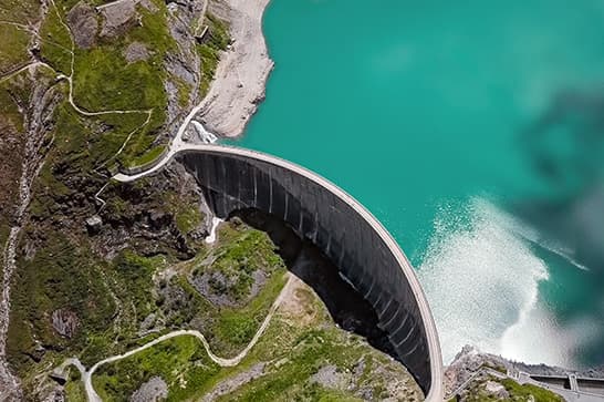 Aerial view of an arc-shaped concrete dam along turquoise water, with green hills, rocky cliffs, and a winding path along the hillside.