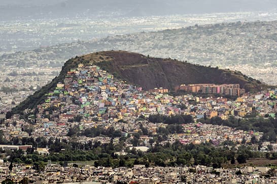 Brightly colored houses densely packed along a hillside, overlooking a sprawling city beneath a hazy sky.