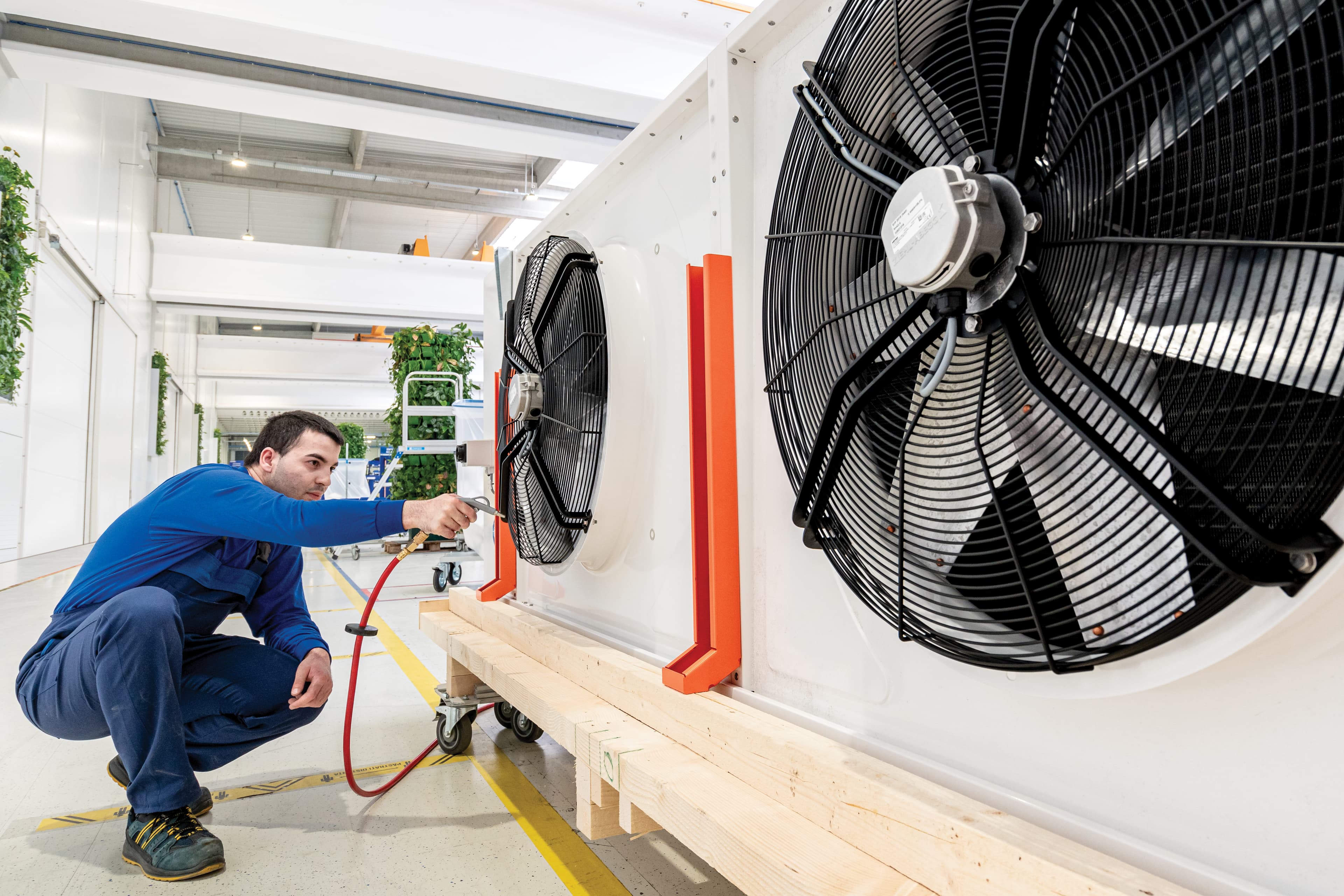 Maintenance worker in blue overalls crouches beside a large industrial cooling unit, adjusting a fan behind a protective guard in a bright workshop.
