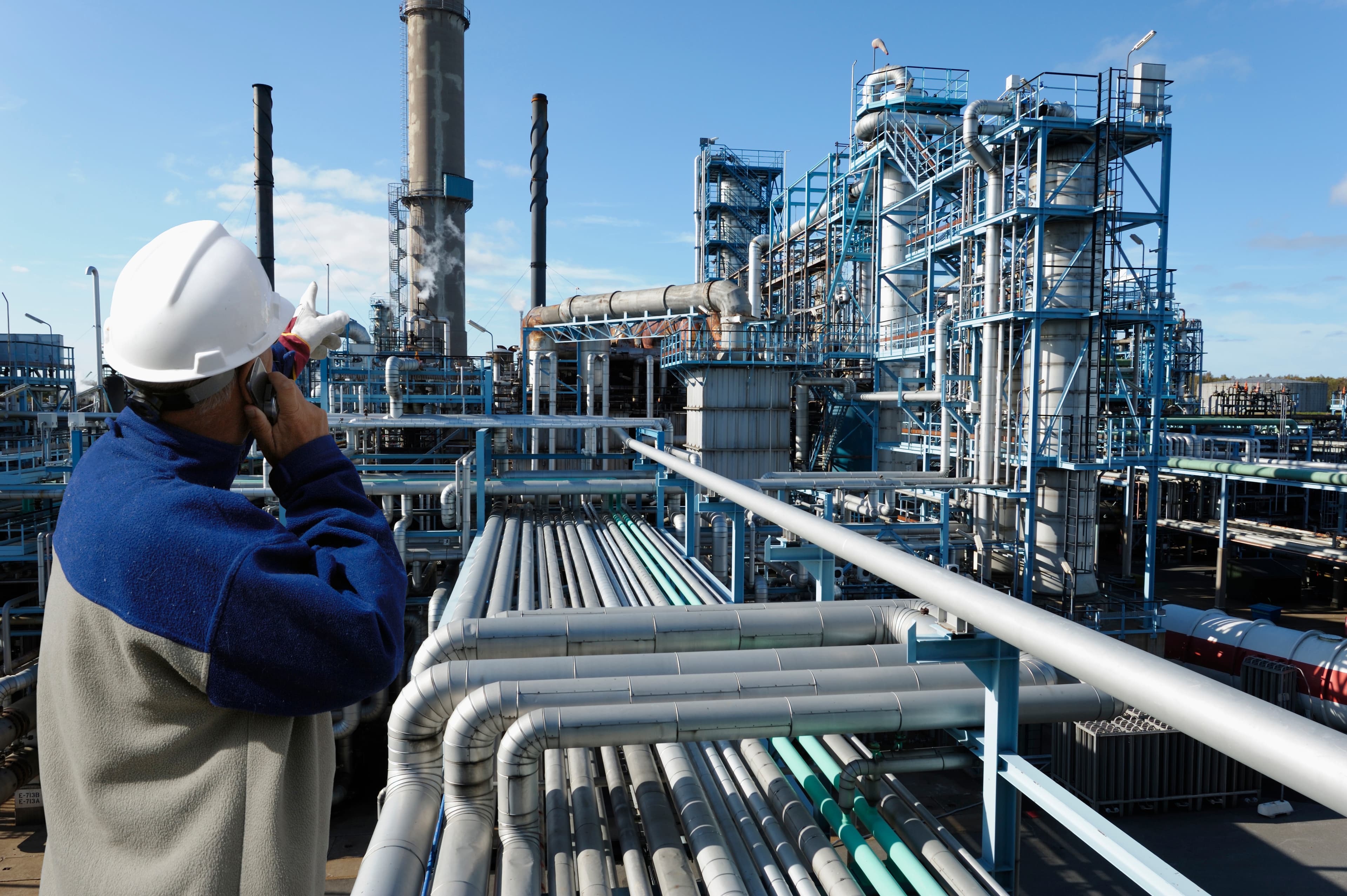 Worker in a white hard hat and blue jacket talks on a phone and points toward an expansive blue industrial refinery with piping and tall stacks.