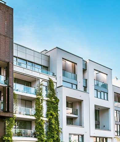 Immeuble résidentiel moderne à façades blanches et gris clair, balcons en verre, vigne sur la façade gauche et ciel bleu vif.