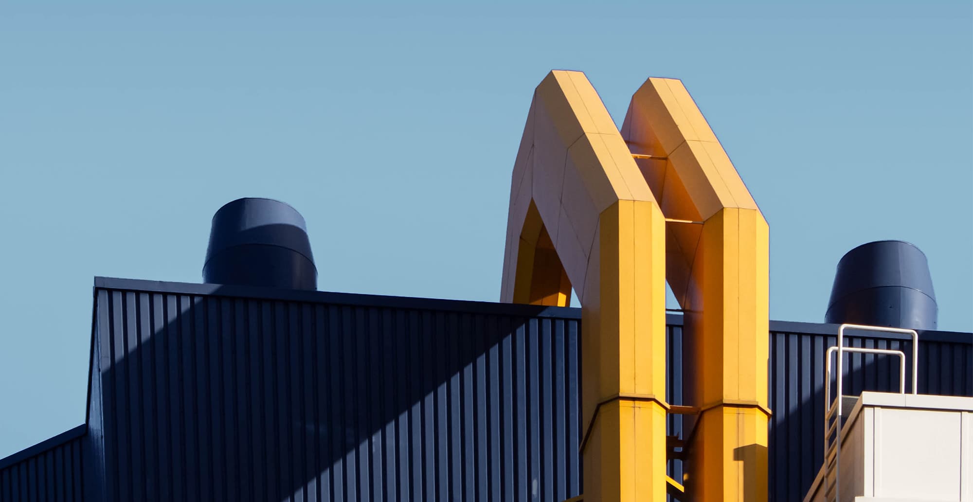 Close-up of a dark blue corrugated roof with a bright yellow double-arch frame and two dark blue vents against a clear blue sky.