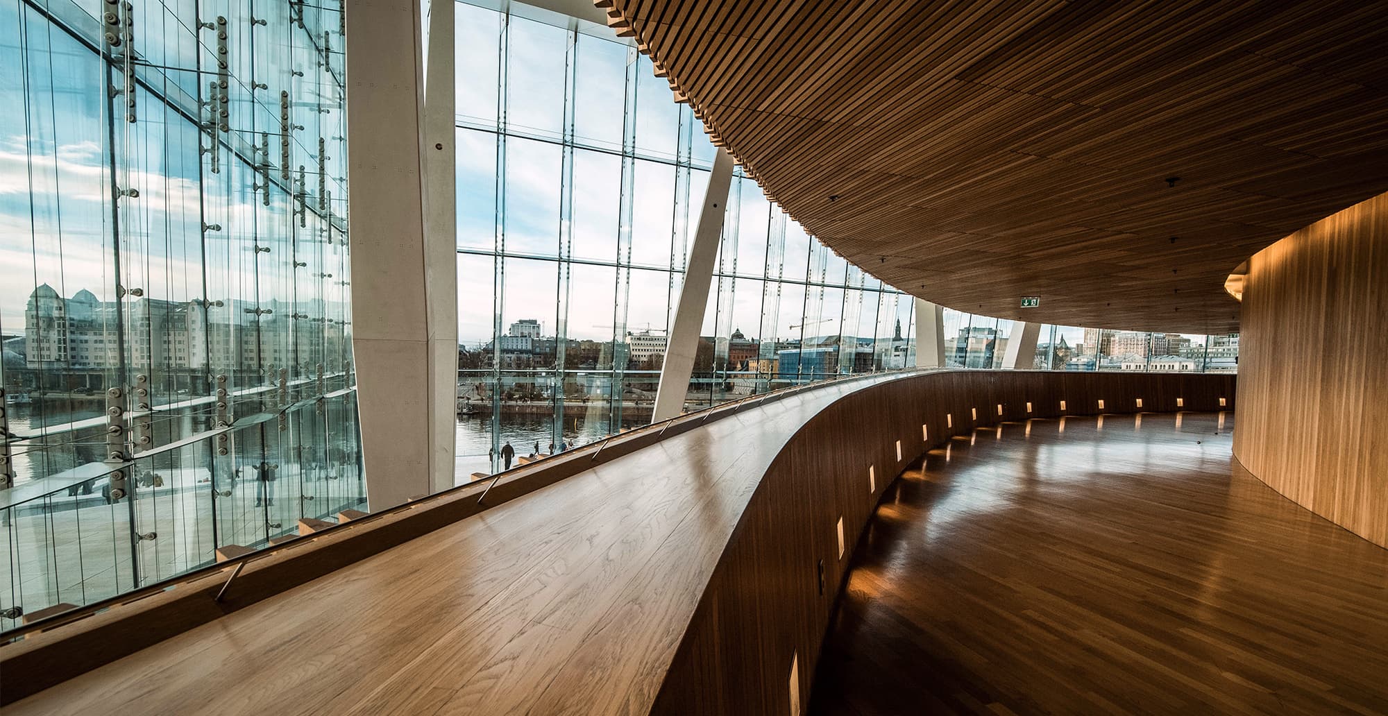 Curved wooden interior walkway with glass walls and white pillars, overlooking a river and cityscape; warm ambient lights along the curved railing.