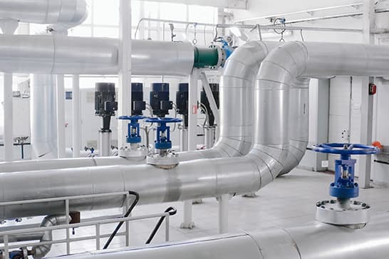 Industrial mechanical room with silver insulated pipes, a large U-shaped duct, blue-handled valves, and wall-mounted control devices.