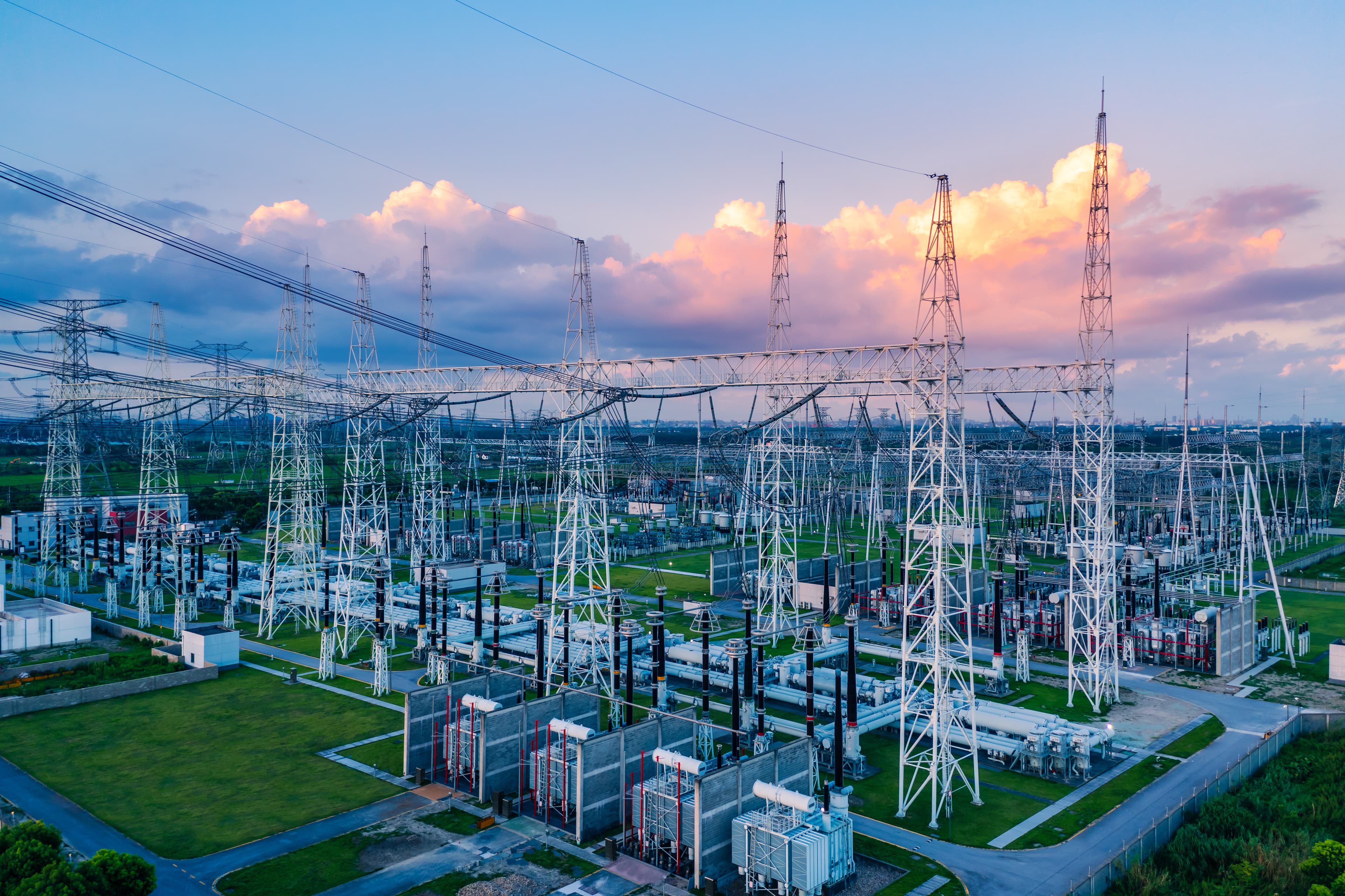 A sprawling electrical substation with white lattice towers, transformers, and cables across green lawns under a pink-orange sunset sky.