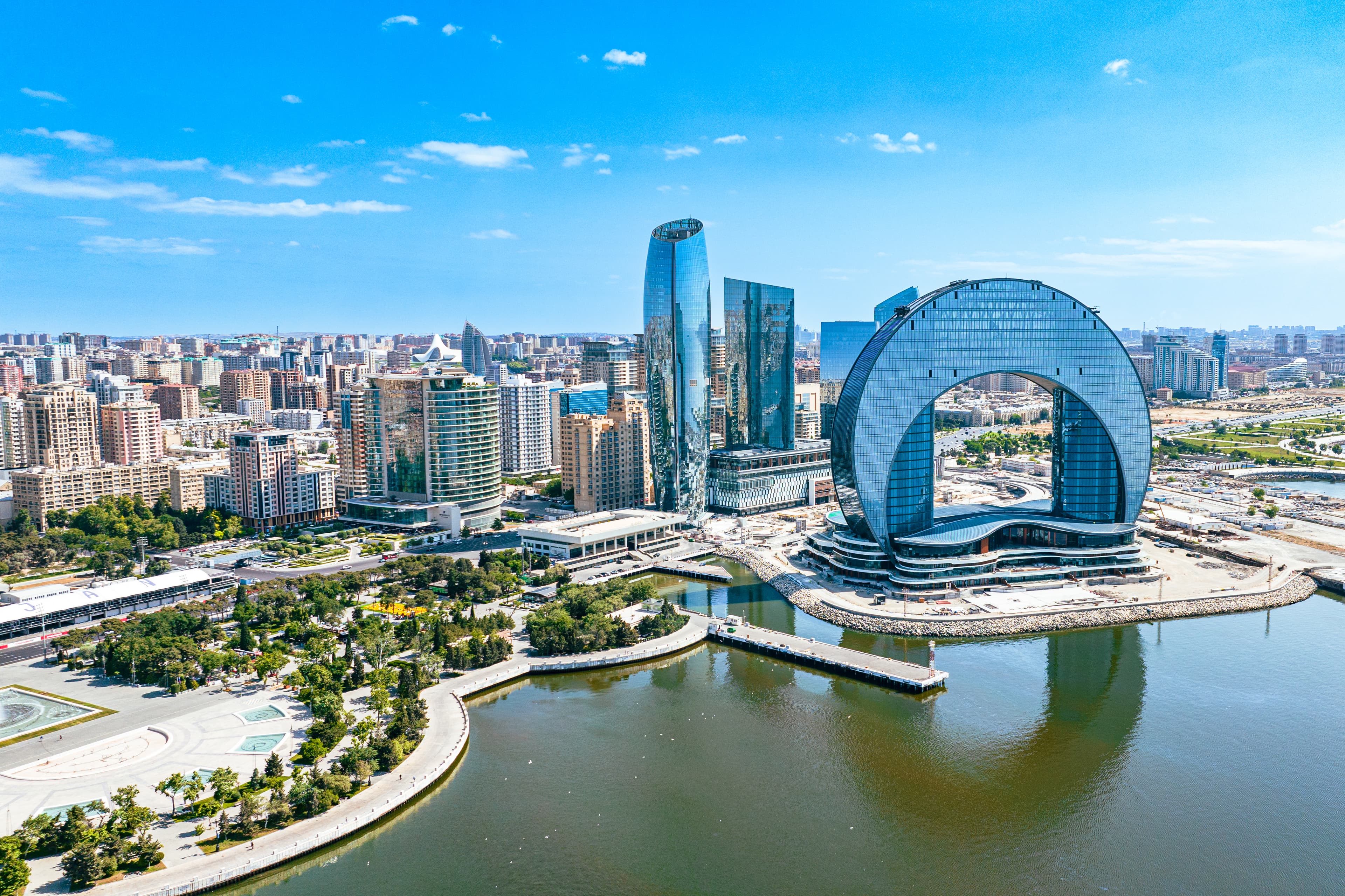 Modern city skyline by a river featuring glass skyscrapers and a large circular arch structure on the waterfront, with a park in the foreground.