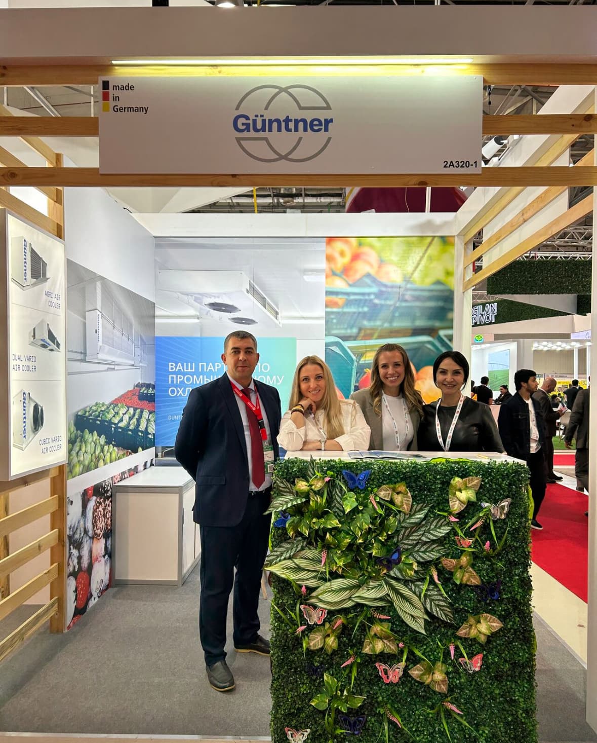 Four people stand at a Günther trade show booth behind a plant-covered counter with butterfly decorations; sign reads Günther, made in Germany.