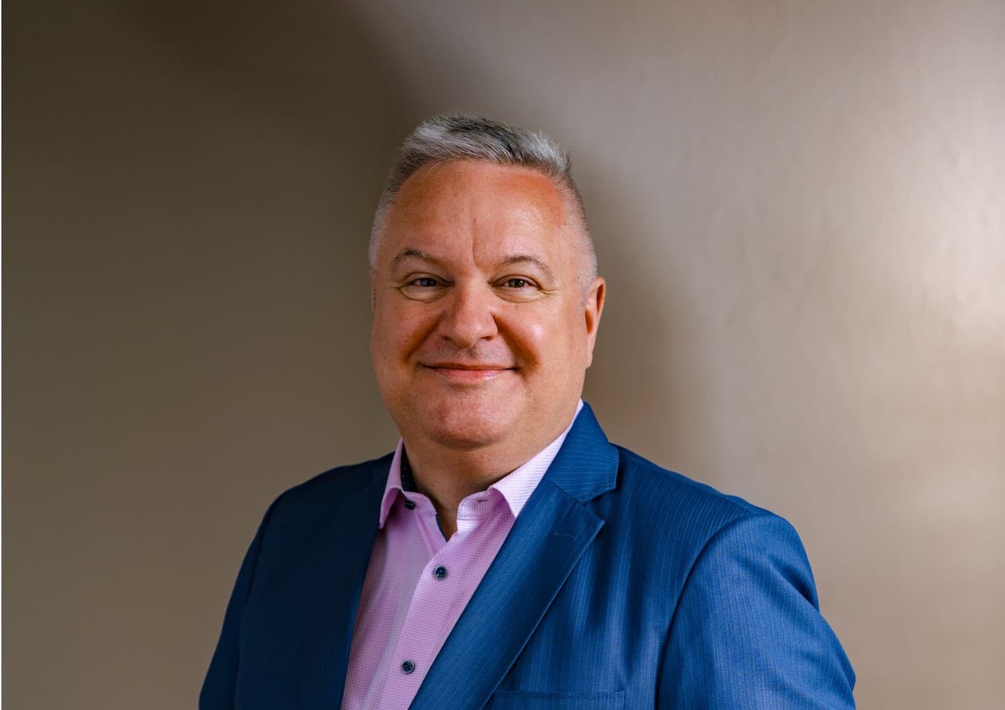 A smiling person with short gray hair wearing a blue suit and pink shirt, standing against a neutral beige background.