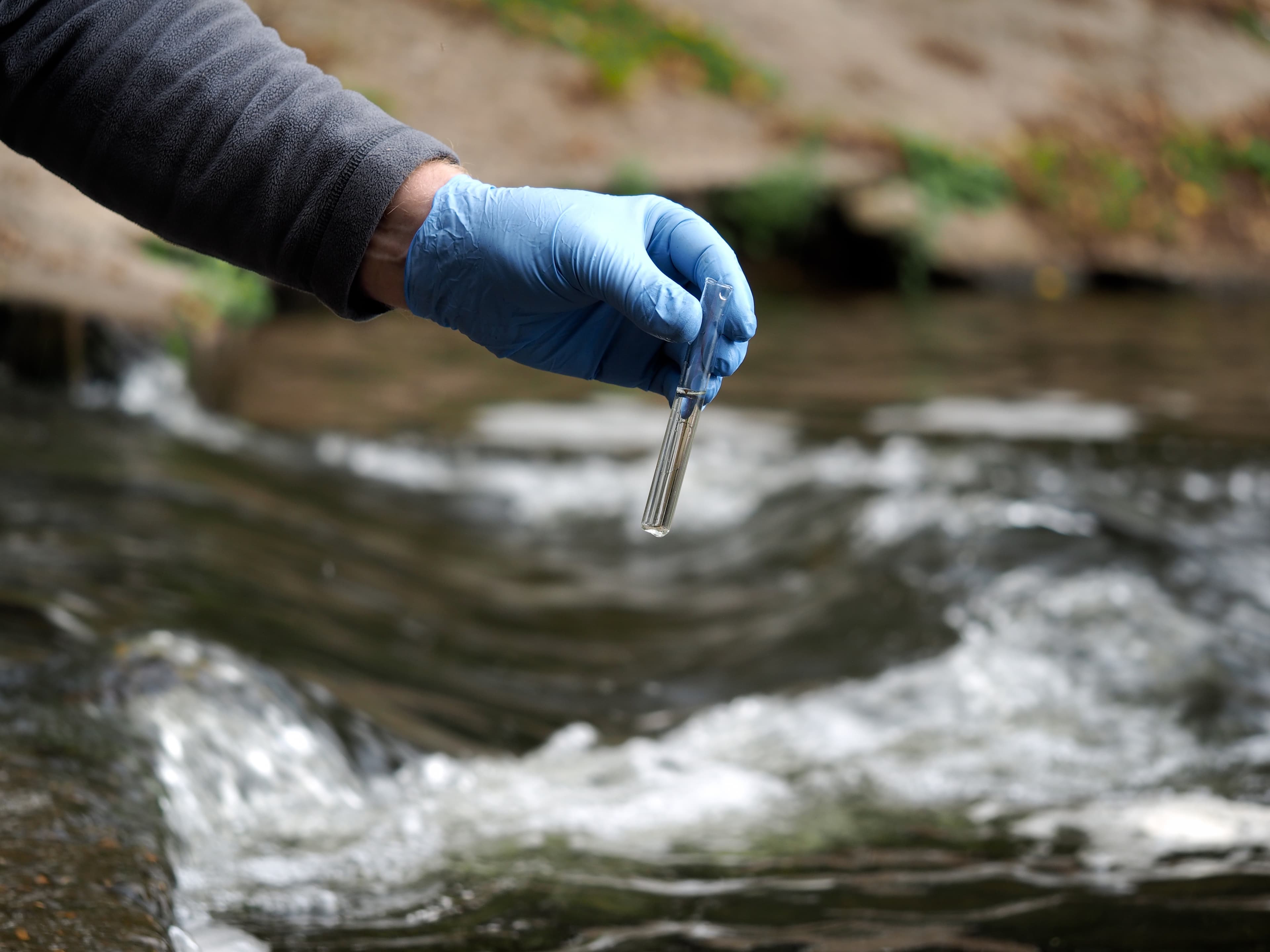 Blue-gloved hand holds a small glass test tube above a flowing stream, with blurred rocks and greenery in the background.