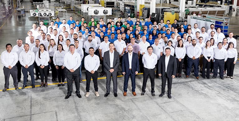 Group photo of factory workers and managers on the factory floor; many wear white shirts and dark pants, with front-row members in suits.