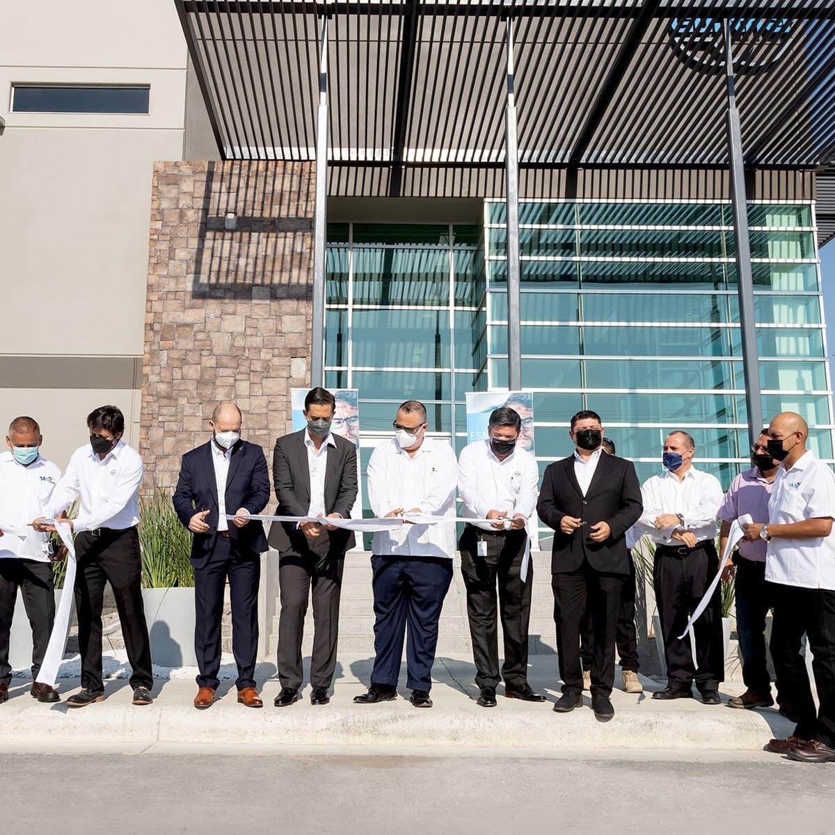 Group of people in business attire and masks cutting a ribbon outside a modern glass-front building with a stone accent wall.