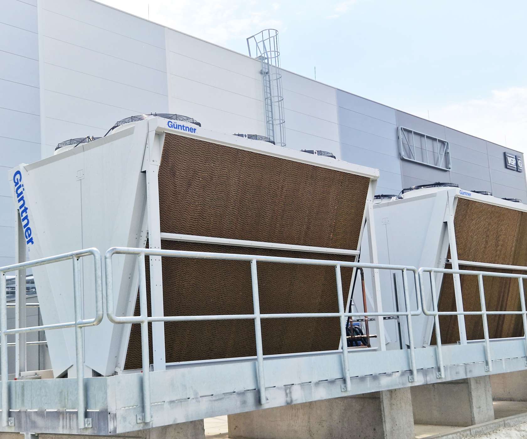 Two large white Guntner cooling units with brown finned panels on a raised platform with railing, beside a modern gray industrial building.