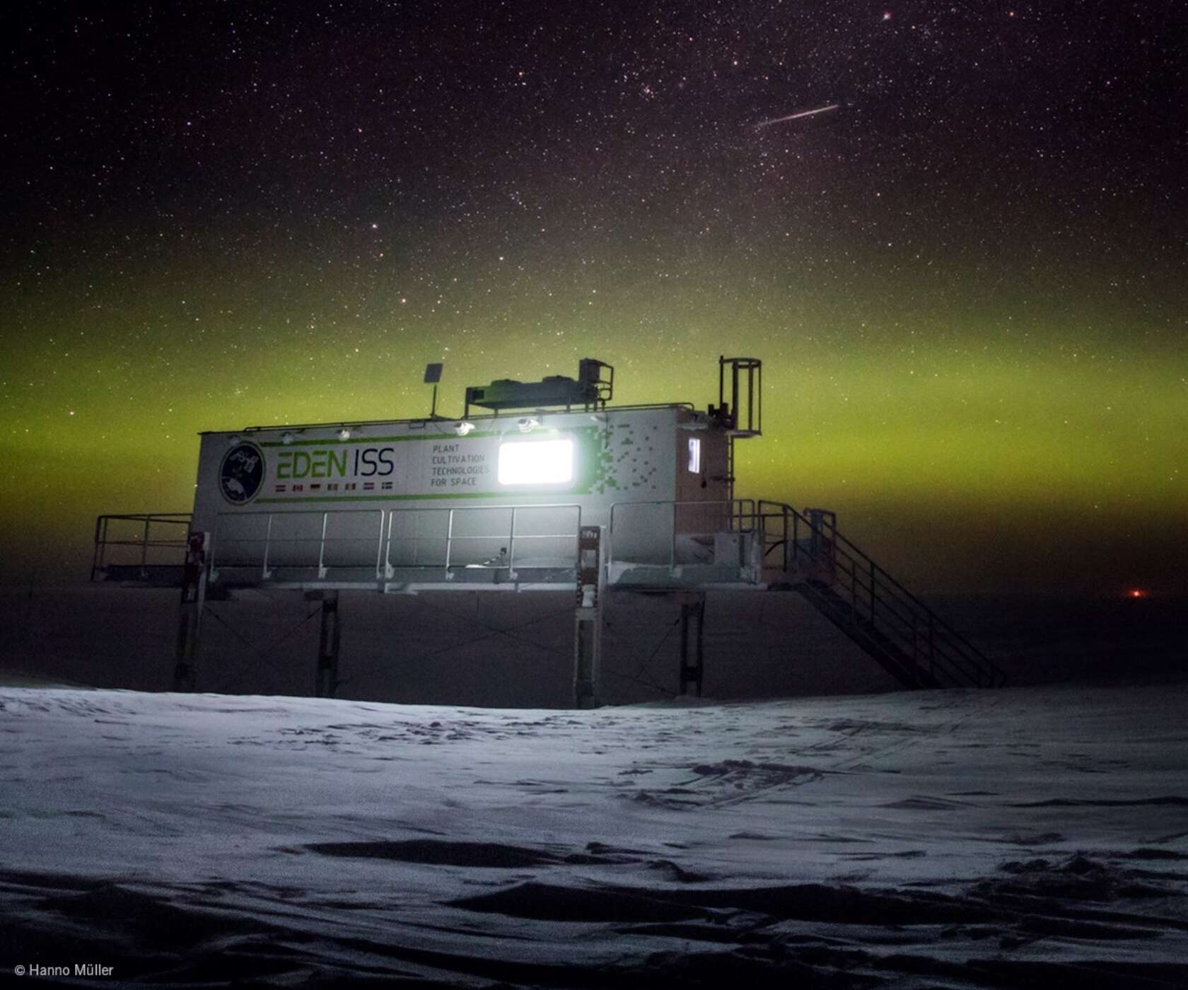 Nighttime view of a raised research module labeled EDEN ISS on stilts above a snow-covered landscape, beneath a green aurora and a starry sky.