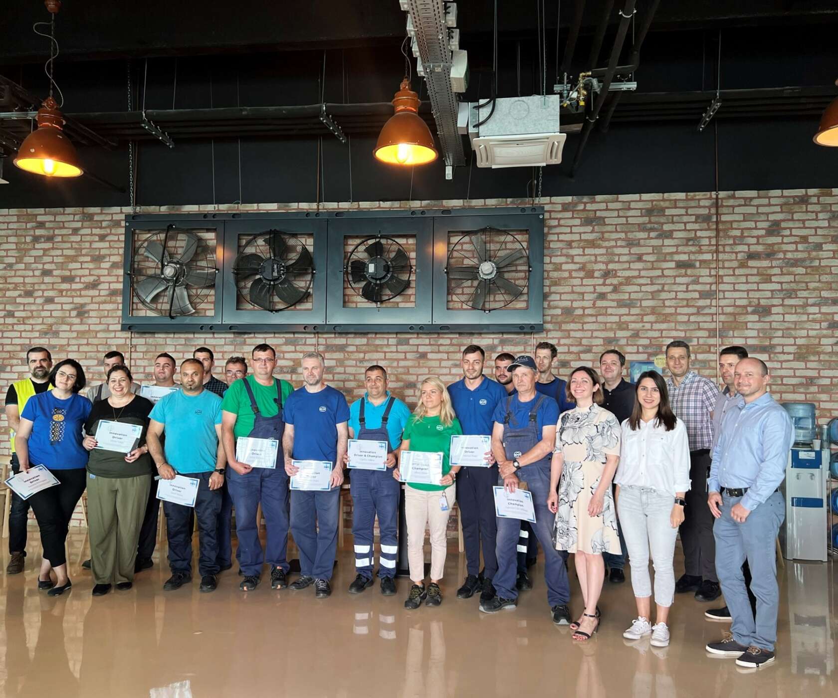 Grupo de personas posando en un taller con paredes de ladrillo, ventiladores industriales y luces naranjas; varios sostienen certificados.
