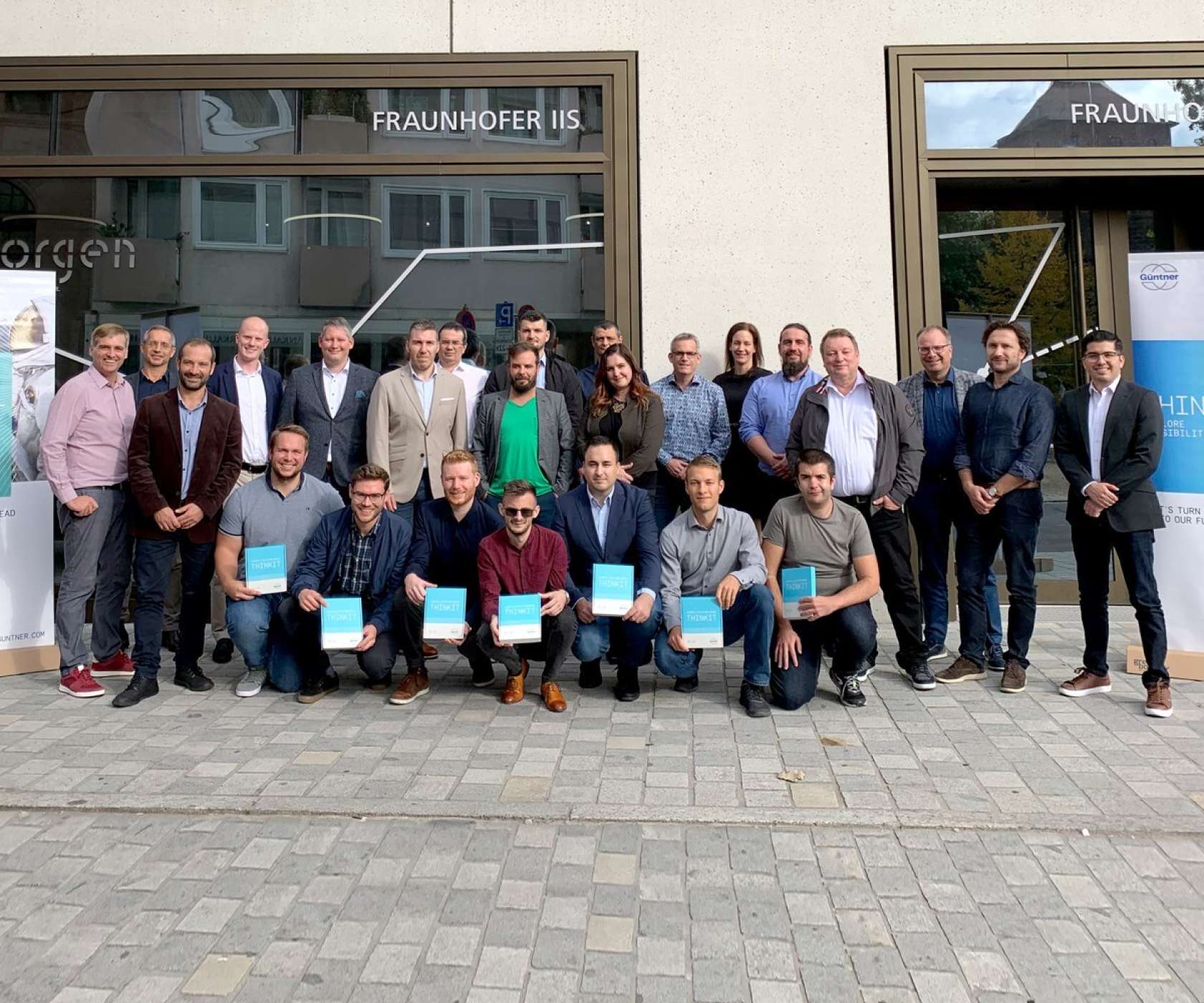 건물 앞에서 여러 사람이 단체 사진을 촬영 중이며, 앞줄이 파란 표지의 책자들을 들고 있다. 배경에 FRAUNHOFER IIS 간판이 보인다.
