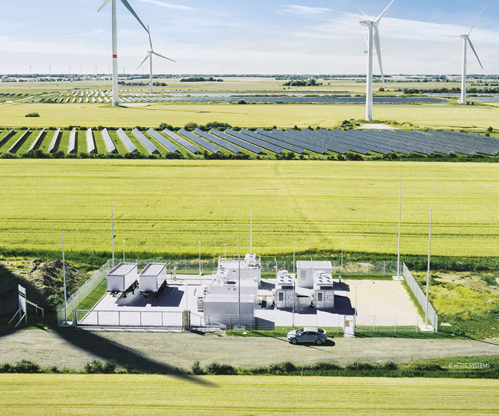 Aerial view of a rural energy site: rows of solar panels, several wind turbines on the horizon, and a fenced utility facility with a car nearby.