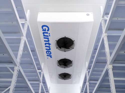 Ceiling-mounted white cooling unit with three circular fans and blue "Guntner" branding, set between white metal racks.