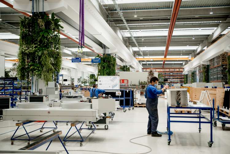 Interior de fabrică modernă, cu plante suspendate și echipamente pe mese; doi angajați în combinezoane albastre lucrează la utilaje.