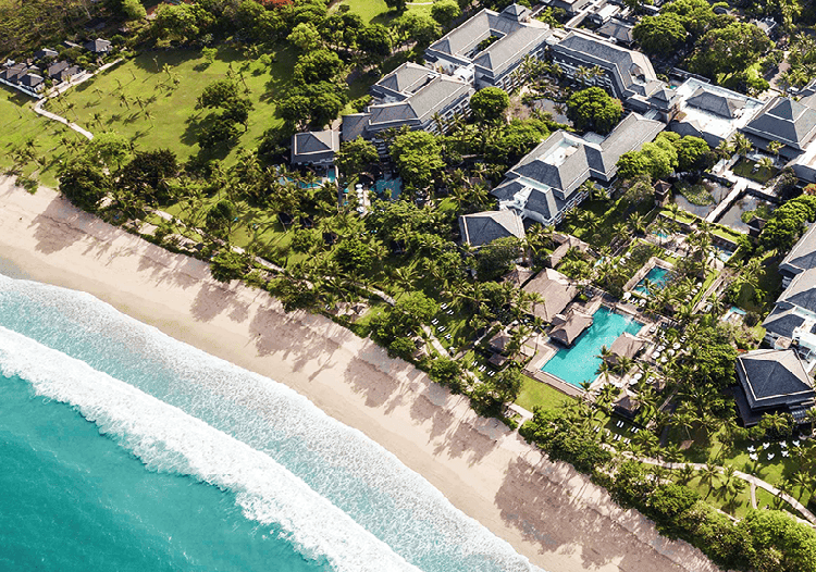 Aerial view of a tropical resort along a sandy beach with turquoise water, palm trees, and clusters of gray-roofed villas surrounding a pool.