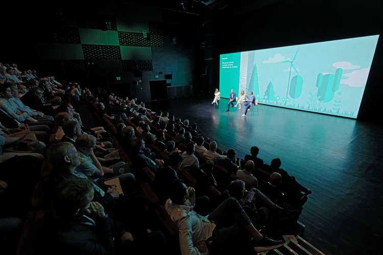 Audience in a dark theater watches four panelists on a lit stage, with a large teal screen showing wind turbines and trees.