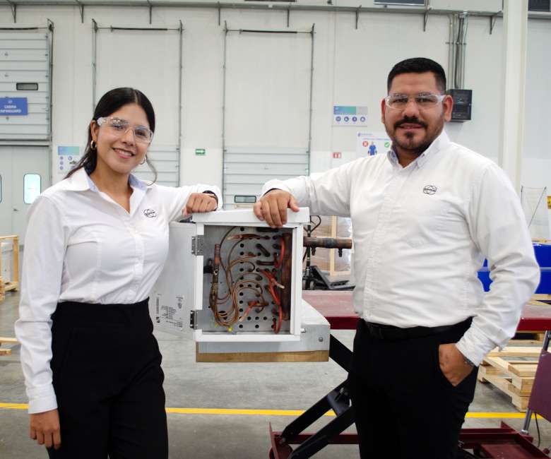 Two people in safety glasses and white shirts stand beside an open electrical cabinet, showing copper wires, in a factory workshop.