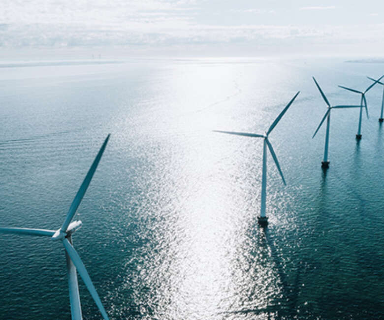 Offshore wind turbines lined up across a calm blue sea, with sunlight reflecting off the water under a pale sky.