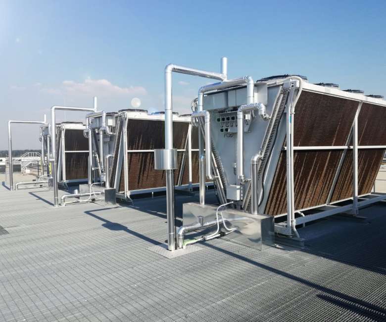 Rooftop scene with multiple large industrial cooling units framed in metal, interconnected silver pipes, and brown heat-exchanger fins under a clear blue sky.